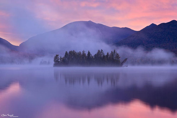 An island on a lake with mountains in the distance during a pink sunrise