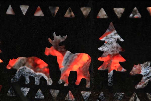 Bright red embers behind a fire grate with bear, moose, and tree shaped openings