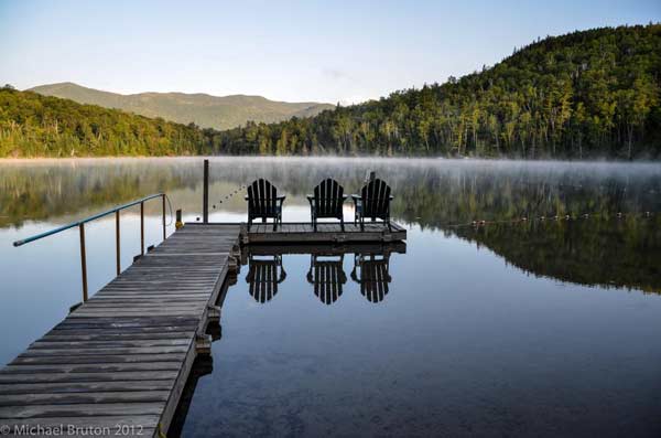 3 Adirondack chairs on a dock over still lake