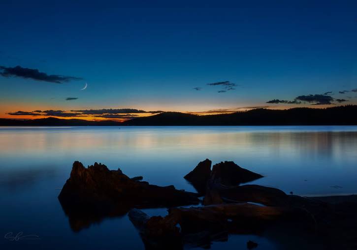 Blue and orange sky with crescent moon over lake