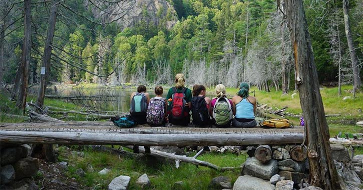 five campers and a counselor sitting on a log in the woods